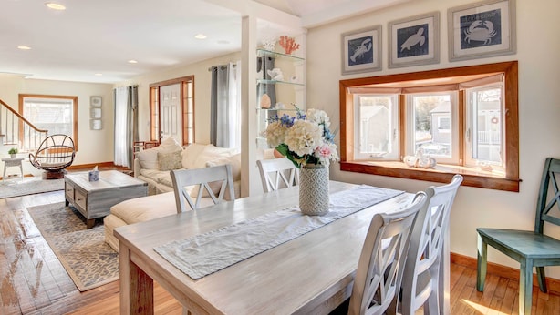 Amplia sala de estar con una mesa de comedor, sillas blancas, una mesa de centro de madera y un jarrón decorativo con flores. Se ve una escalera que conduce a otro nivel, y una ventana con cortinas.
