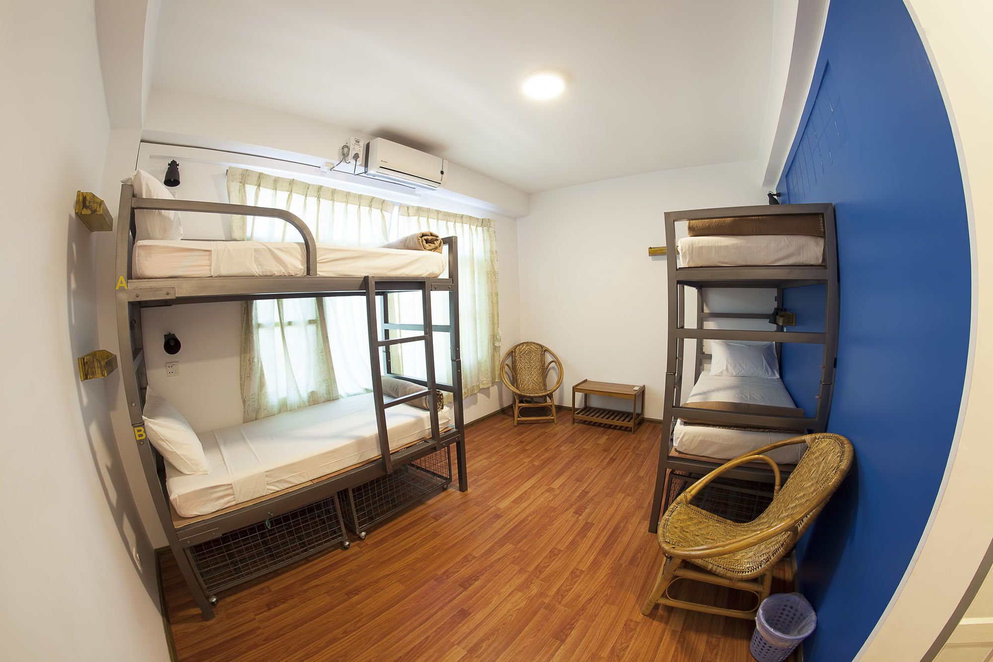 A dormitory room with bunk beds, a standing heater, a wicker chair, and a small table.