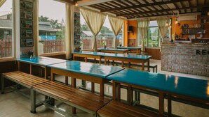 A restaurant with wooden tables and benches, large windows with curtains, and a brick wall with a sign. - Ostello Bello Nyaung Shwe (Nyaungshwe)