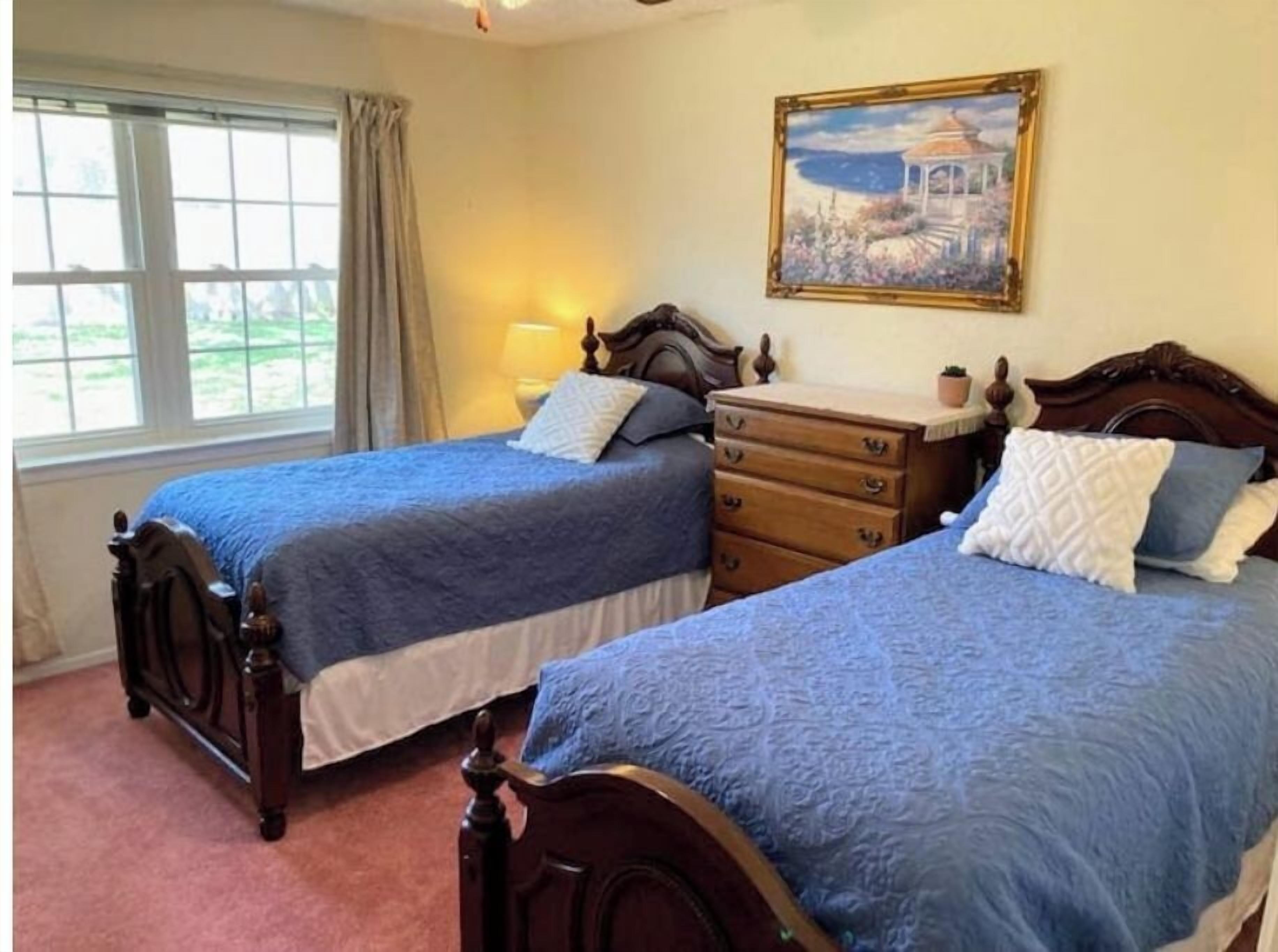 A room with two wooden beds with blue bedding, a wooden dresser, a painting on the wall, and a window with curtains.