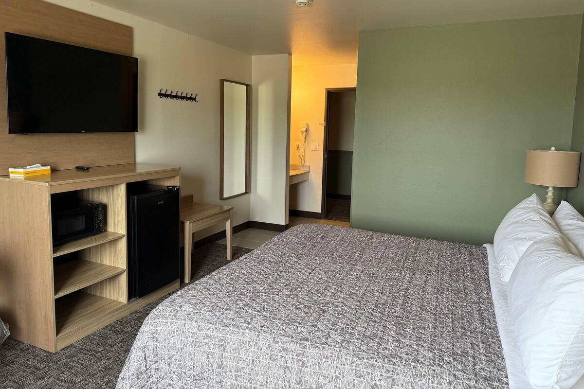 A hotel room with a bed, a TV mounted on the wall, a small desk with a microwave, and a bathroom visible through an open door.