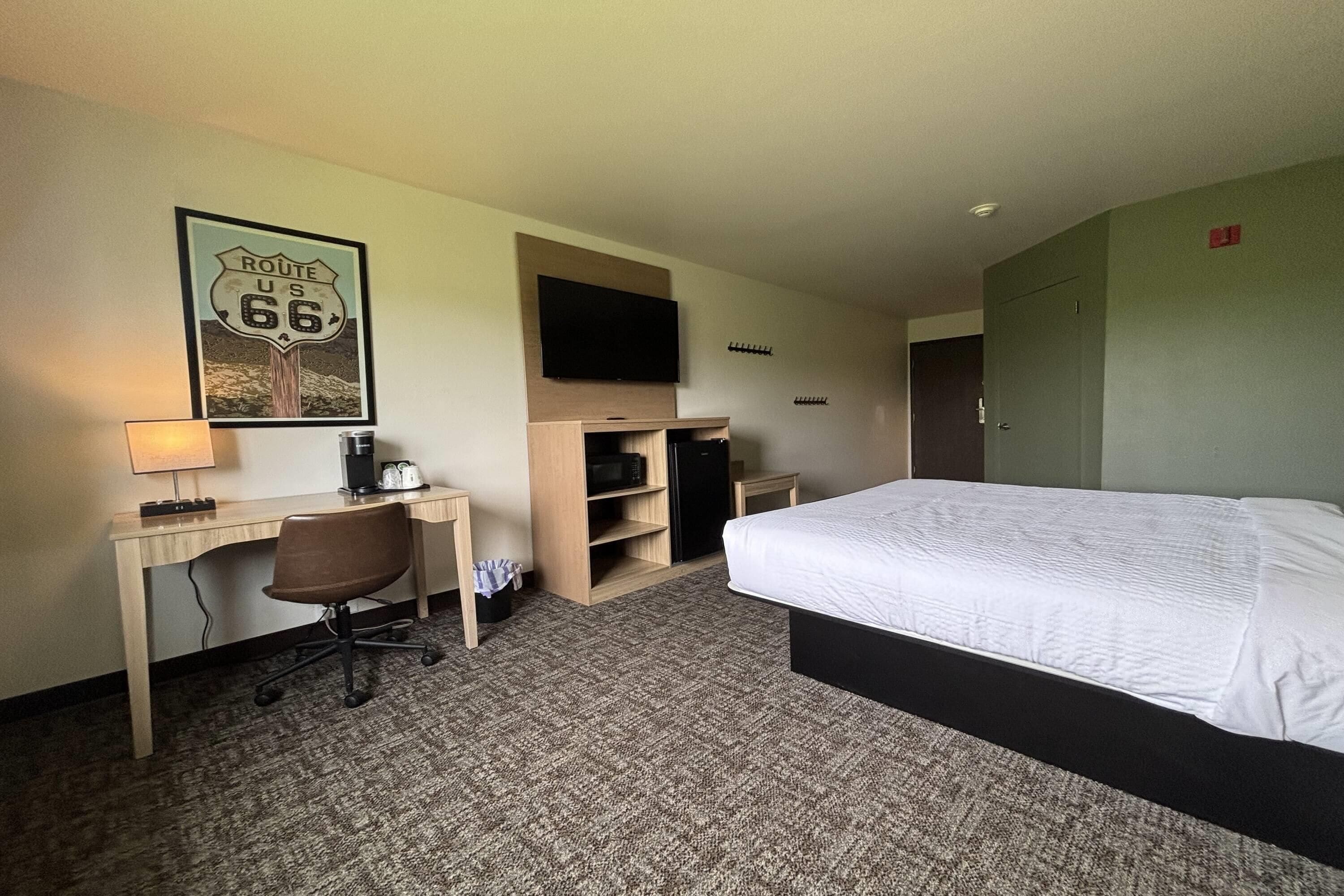 A hotel room with a bed, desk, chair, TV, and a framed picture of Route 66.