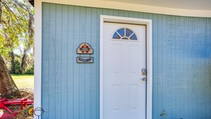 Ein Haus mit weißer Tür, einem Willkommensschild und einer kleinen Veranda mit einem roten Stuhl und einem blauen Werkzeug.