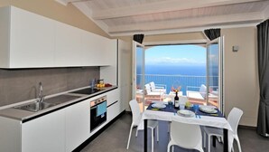 A compact kitchen with a sink, stove, and white cabinets. A dining area with a table and chairs is visible, overlooking a balcony with a view of the sea.