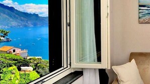 A room with a large window overlooking a coastal landscape, a bed, and a painting on the wall.