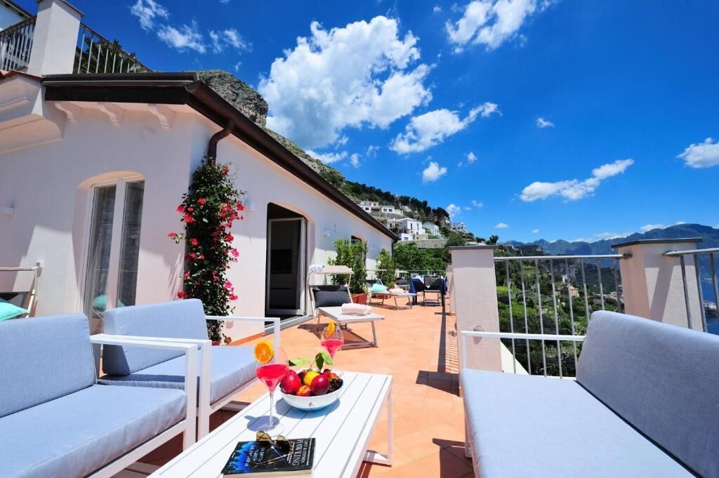 A terrace with white furniture, a table with fruit and drinks, and a view of a hillside with buildings.