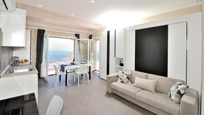 A modern hotel room with a kitchenette, a dining area with a table and chairs, a sofa, and a large window offering a view.