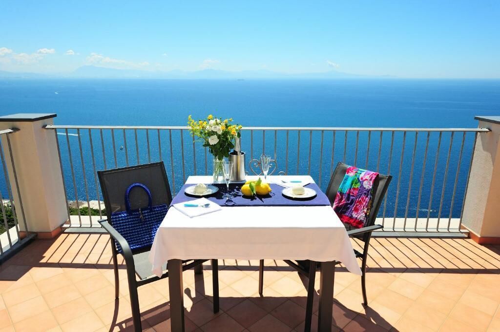Una mesa en un balcón con vistas al mar, un florero con flores y un vaso de agua.