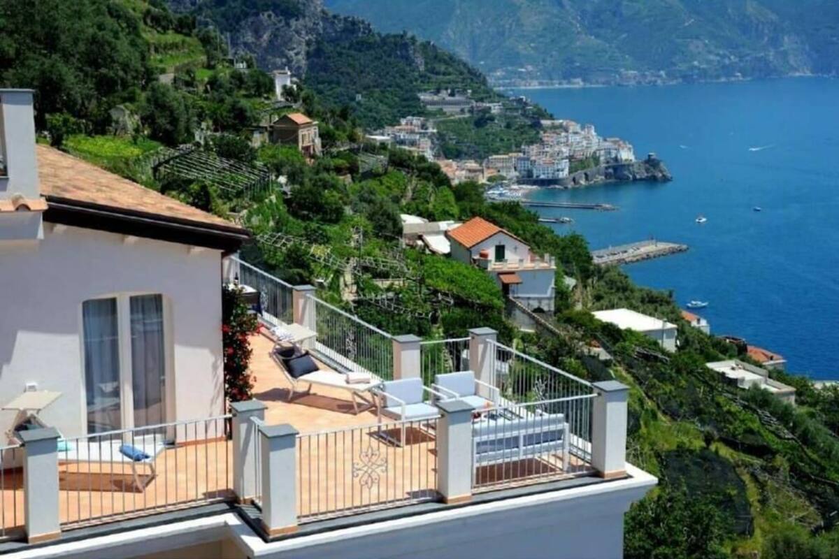 Um imóvel costeiro com varanda e vista para o mar, área de jardim com mobiliário externo e paisagem montanhosa ao fundo.