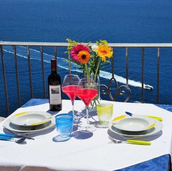 A table set with a bottle of wine, two martini glasses with red liquid, a vase with flowers, and plates with cutlery on a balcony overlooking the sea. - Amalfi Blu Retreat  (Amalfi)