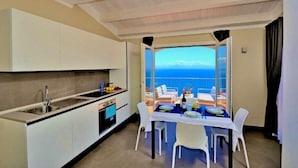 A compact kitchen with a view of the sea, a dining area with a table and chairs, and a balcony with a table set for two.