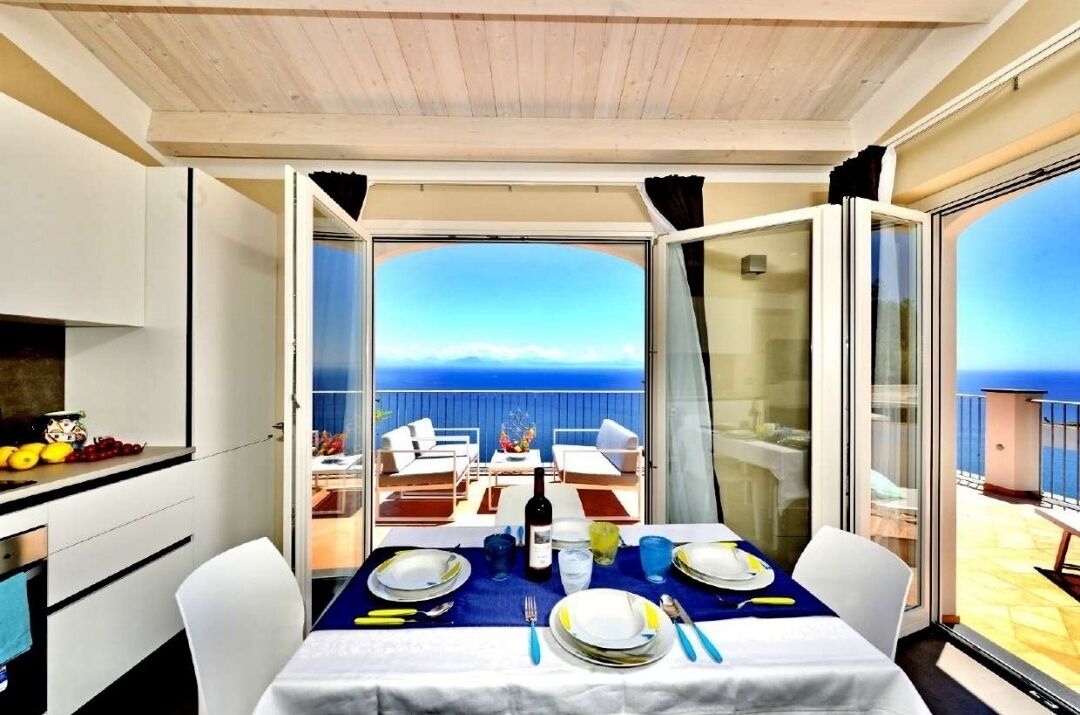A dining area with a table set for two, overlooking a balcony with a view of the sea.