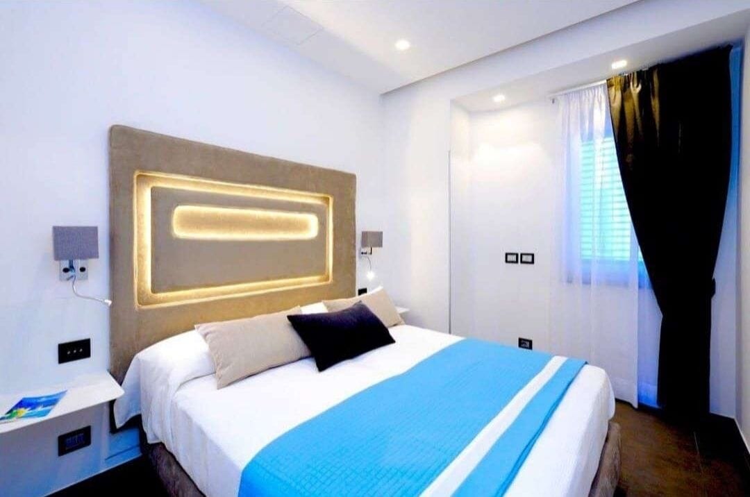 Camera d'albergo moderna con un letto grande, comodini, una testiera con illuminazione integrata, una finestra con tende e un pannello di controllo a parete.