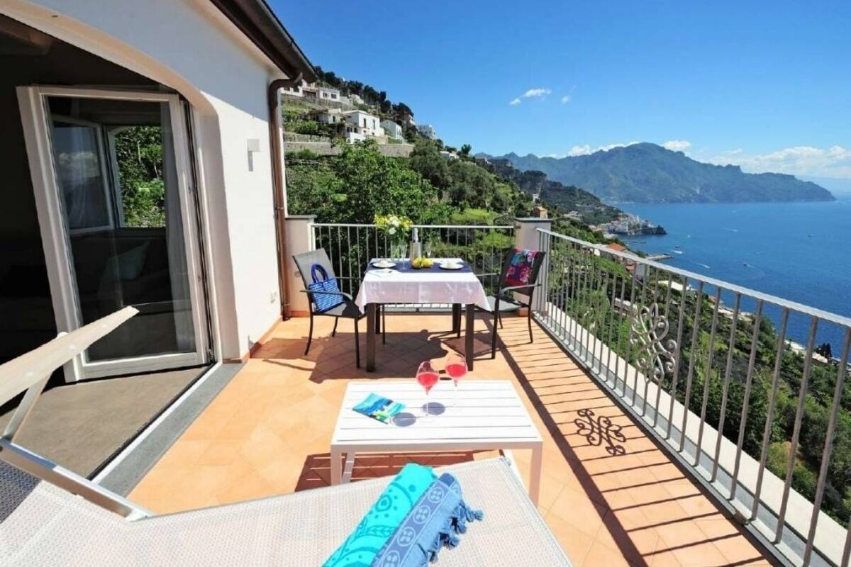 Uma varanda com mesa e cadeiras, vista para o mar e paisagem montanhosa.