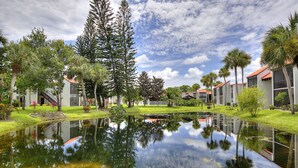 A serene courtyard with a pond reflecting trees and buildings, surrounded by well-maintained lawns and palm trees. - Cozy Condo at Village Brooks (Sarasota)