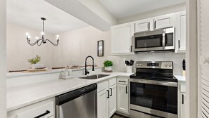 A modern kitchen with white cabinets, stainless steel appliances, a dishwasher, and a black chandelier. - Cozy Condo at Village Brooks (Sarasota)