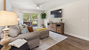 A living room with a sofa, a coffee table, a lamp, a television, and a wooden cabinet. - Cozy Condo at Village Brooks (Sarasota)