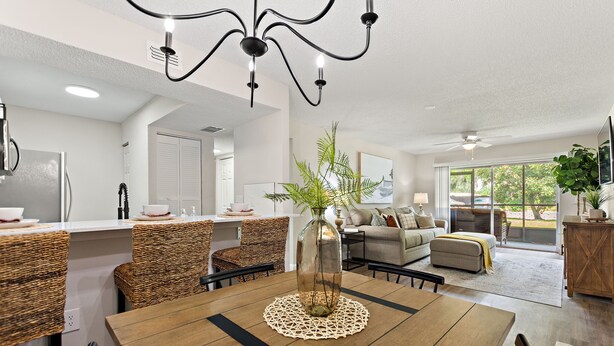 A modern kitchen with a central island, dining area with a wooden table and wicker chairs, and a living room with a sofa and a large window. - Cozy Condo at Village Brooks (Sarasota)