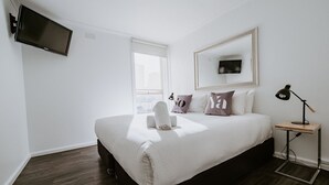 A modern hotel room with a large bed, a flat-screen TV mounted on the wall, a bedside table with a lamp, and a window providing a city view. - Soya Apartment Hotel (Melbourne)