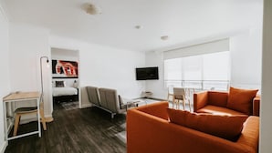 A modern hotel room with a wooden floor, a sofa, a desk with a chair, a bed, a TV, and a window with a view. - Soya Apartment Hotel (Melbourne)