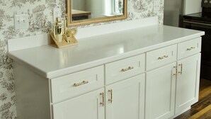 A white vanity with a gold-framed mirror, a white countertop, and a wooden floor.