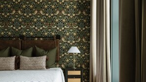 A bedroom with a patterned wallpaper, a bed with a patterned bedspread, a nightstand with a lamp, and a window with curtains.