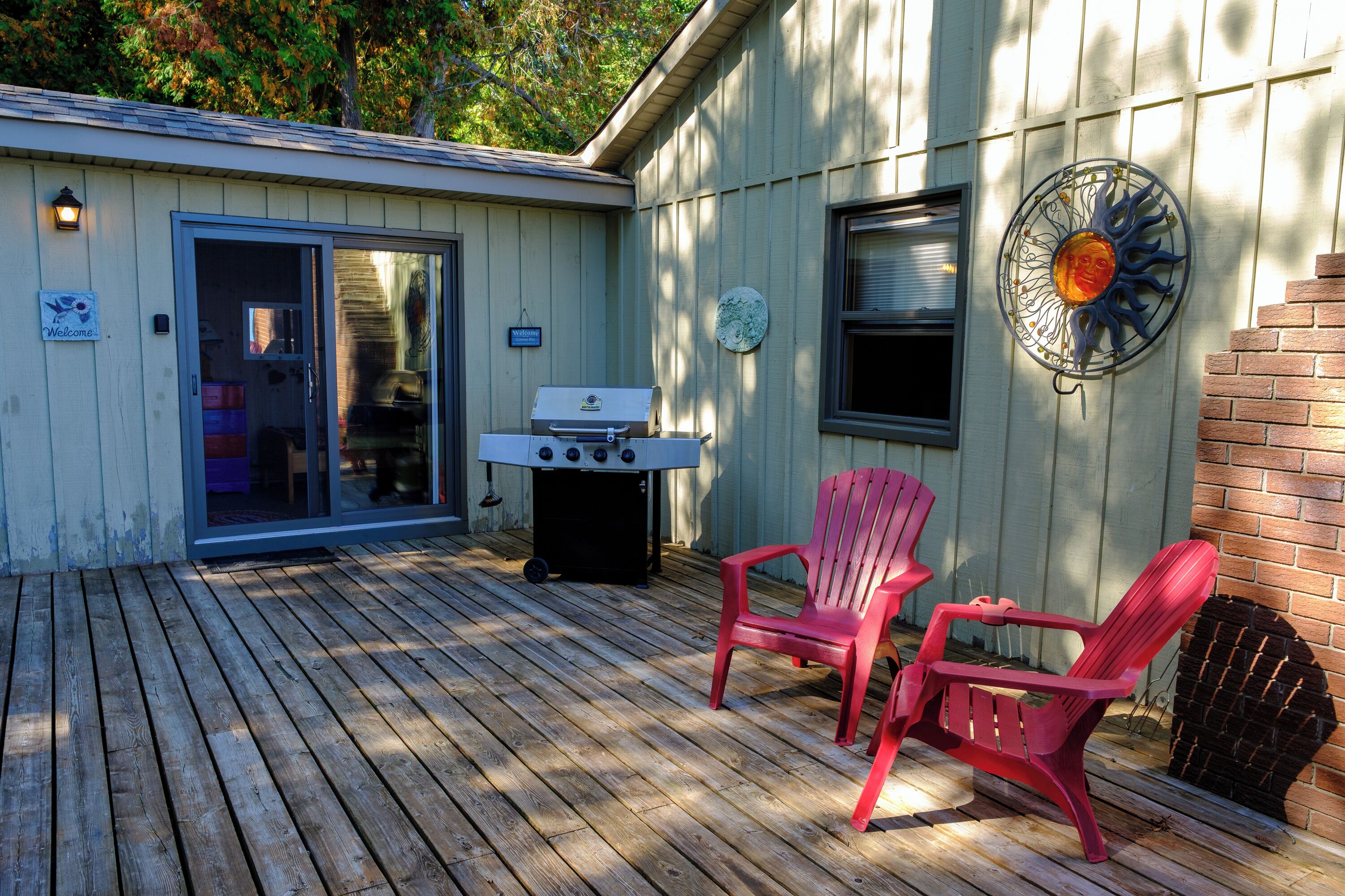 A wooden deck with a grill, two red chairs, and a wall-mounted decorative piece.