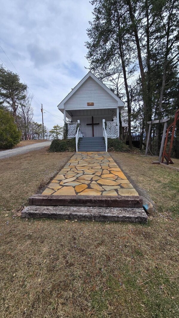 A small white chapel with a cross at the entrance, a stone pathway leading to it, and a wooden fence surrounding the property.
