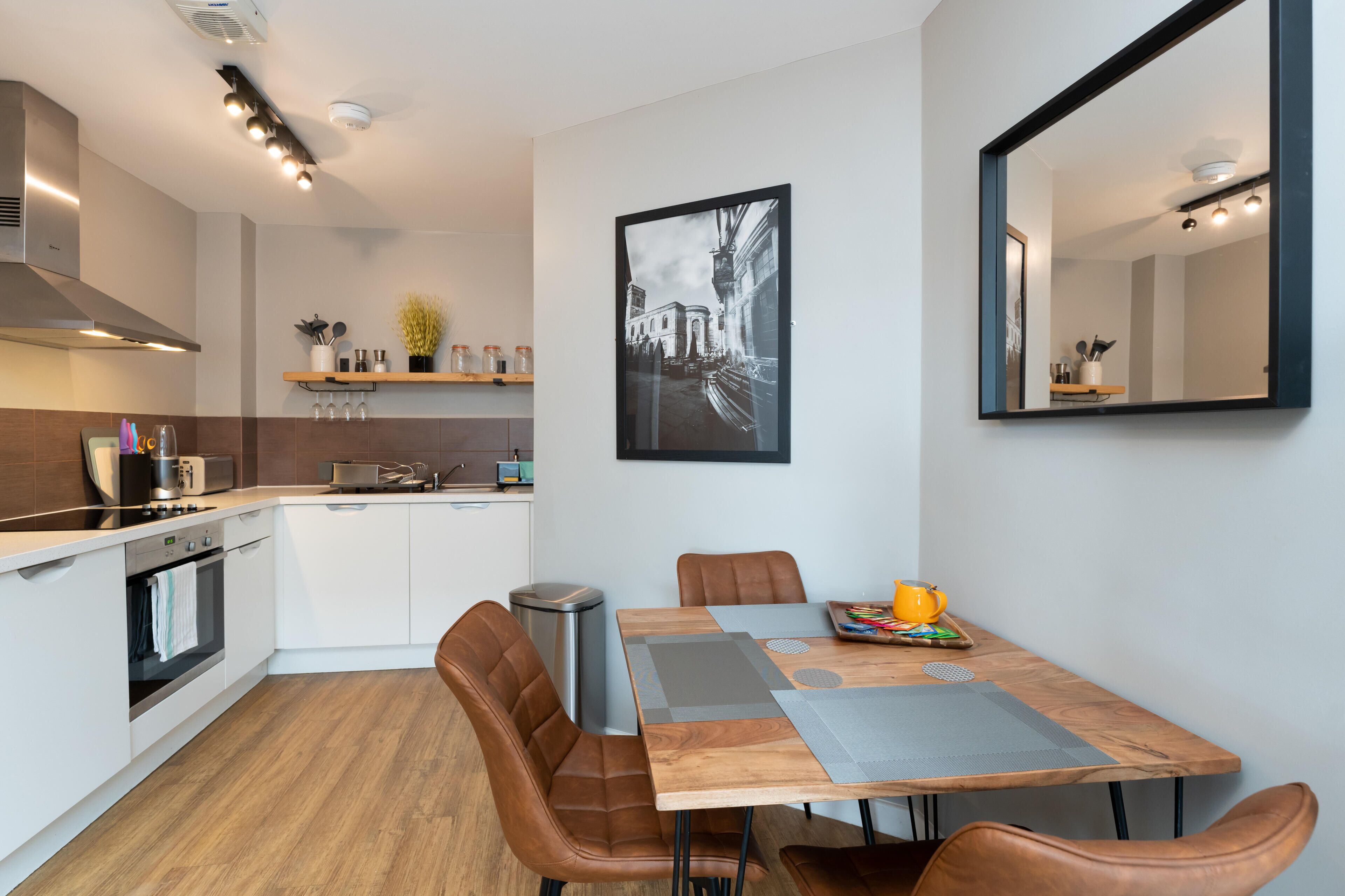 Une cuisine moderne avec des armoires blanches, un four encastré et une hotte de cuisine en acier inoxydable. On y trouve un coin repas avec une table en bois, des chaises en cuir marron et un grand miroir au mur.