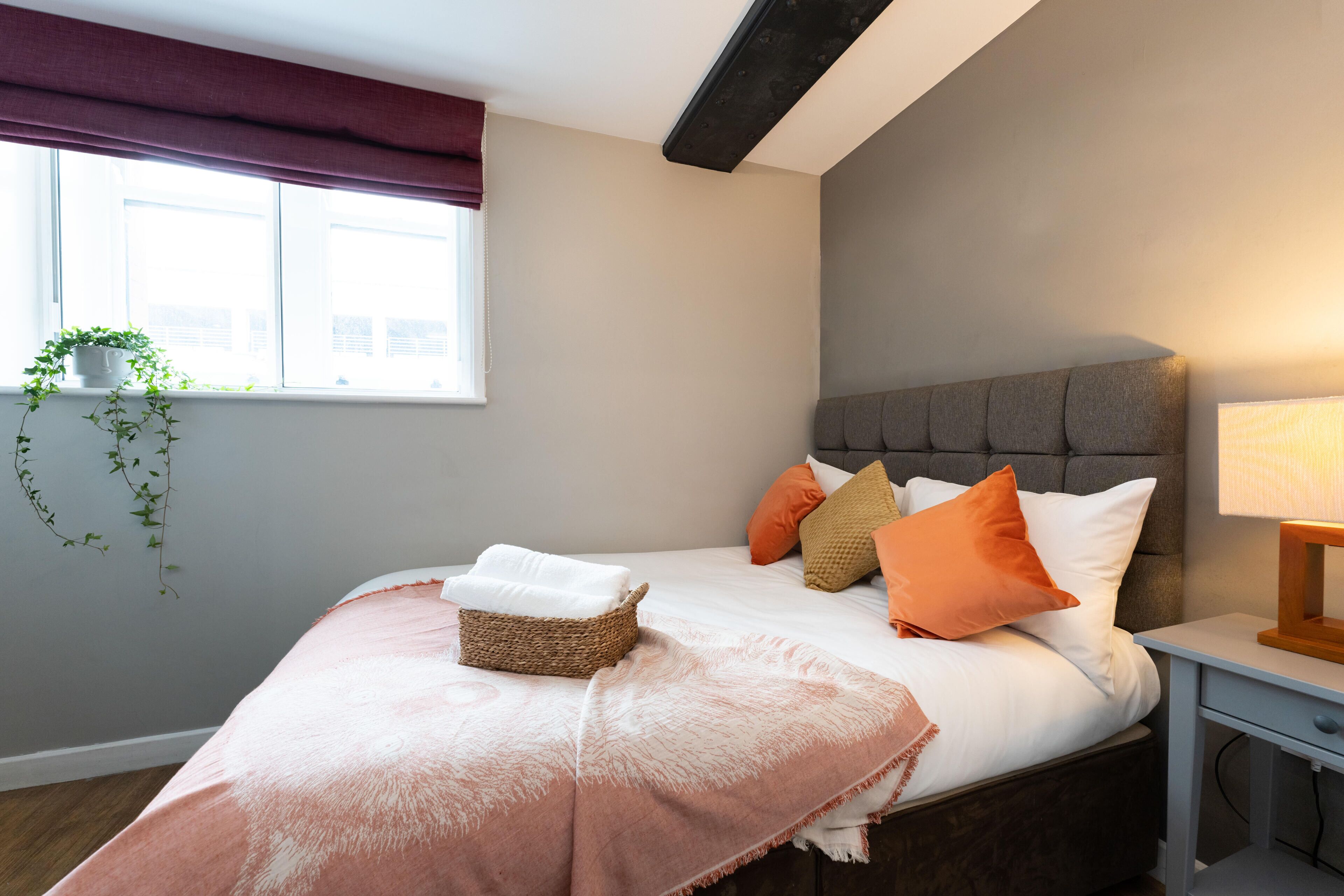 A neatly made bed with a basket of folded towels, a bedside table with a lamp, and a window with a plant.