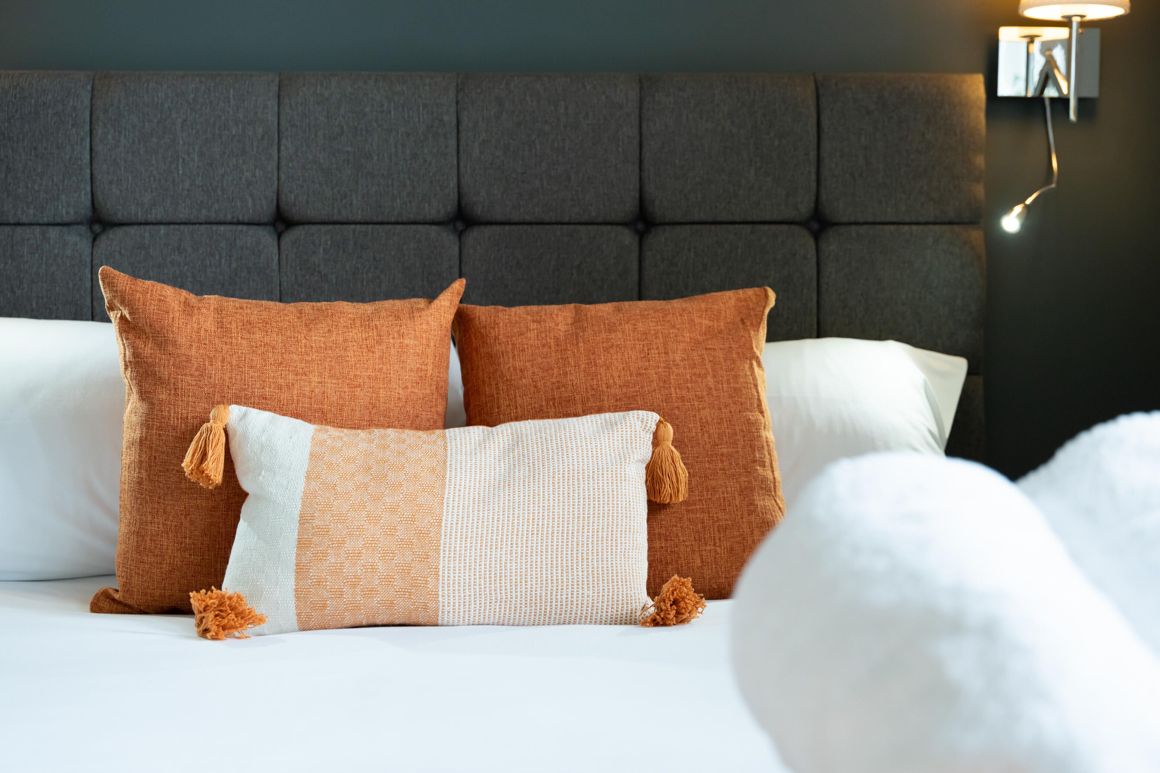A bed with a tufted headboard, two brown pillows, a white pillow, and a white and orange patterned pillow.