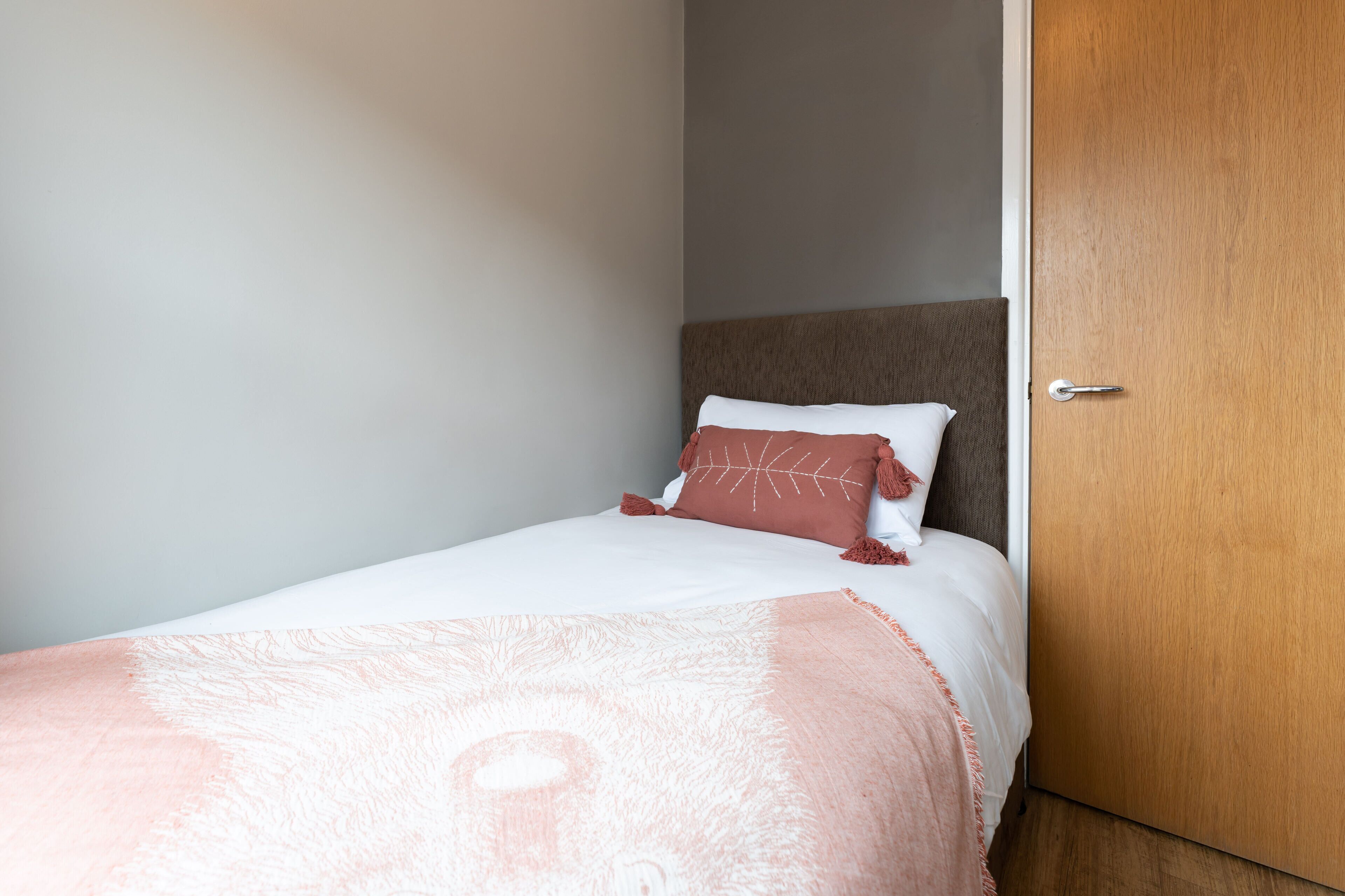 A neatly made bed with a patterned blanket and two pillows, a wooden door, and a plain wall.
