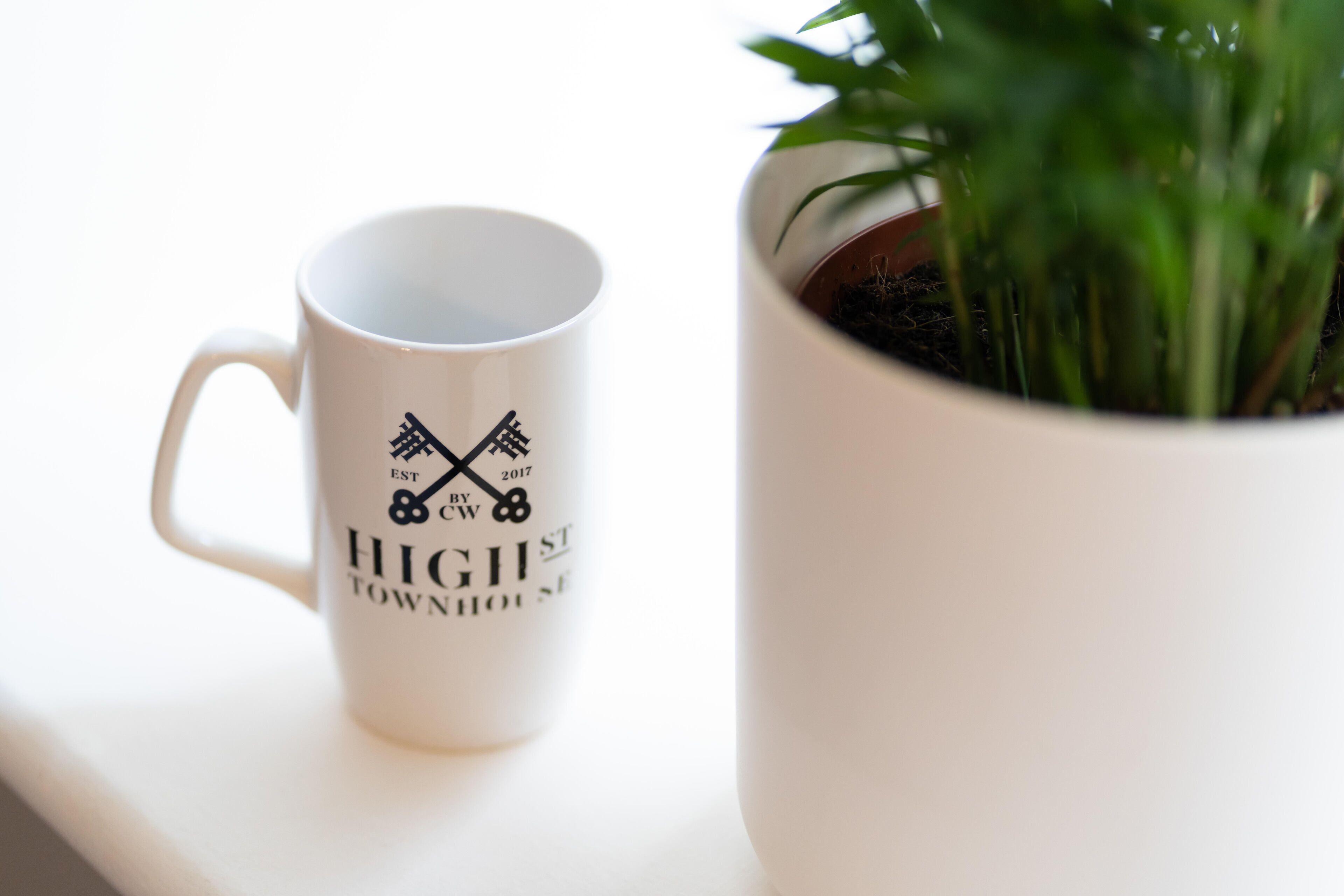 Uma caneca branca com texto preto e um logotipo, ao lado de uma panela branca com plantas verdes.