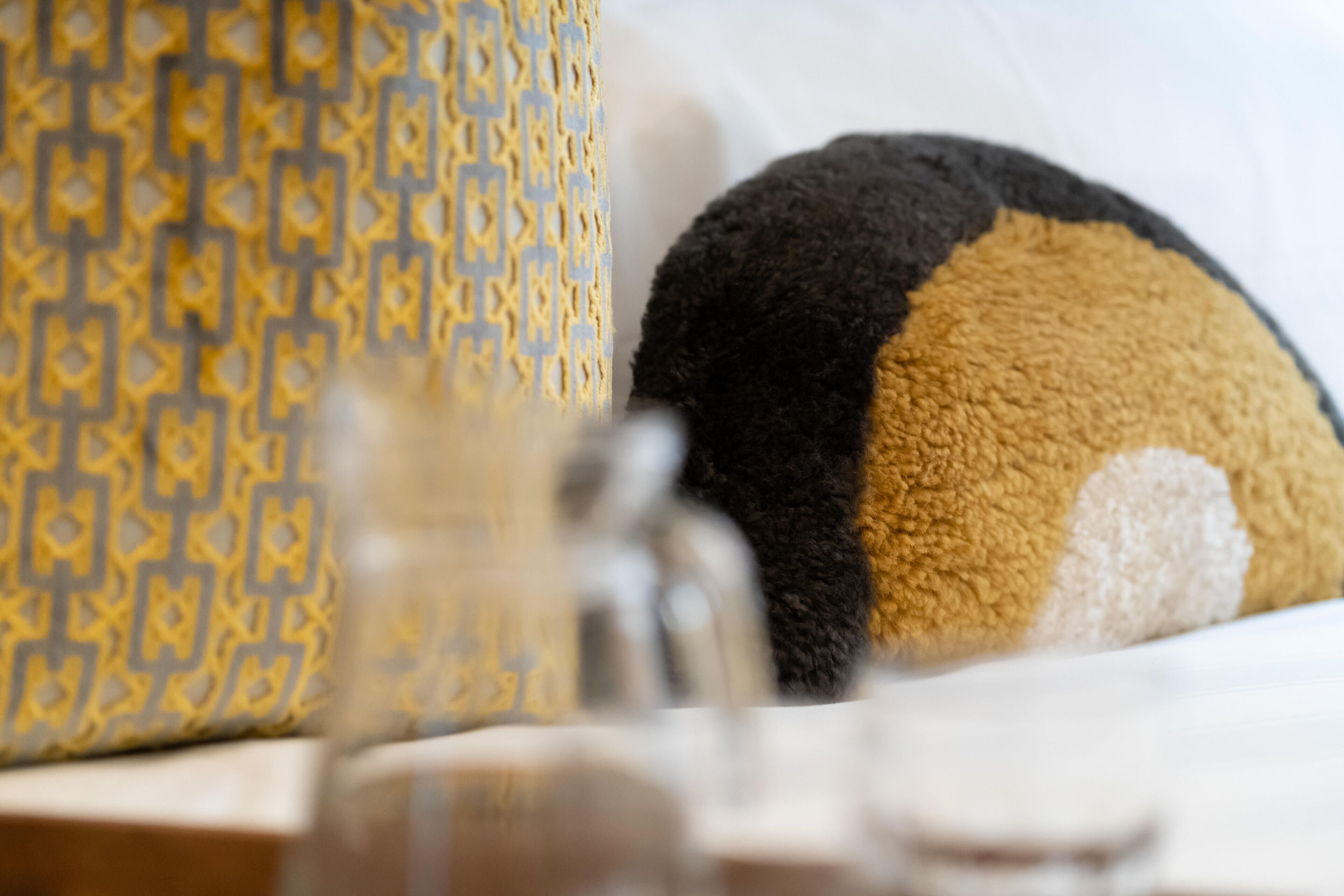 Un peluche que se asemeja a un oso, un cojín amarillo y azul con estampado, y un objeto de vidrio transparente, probablemente una botella.