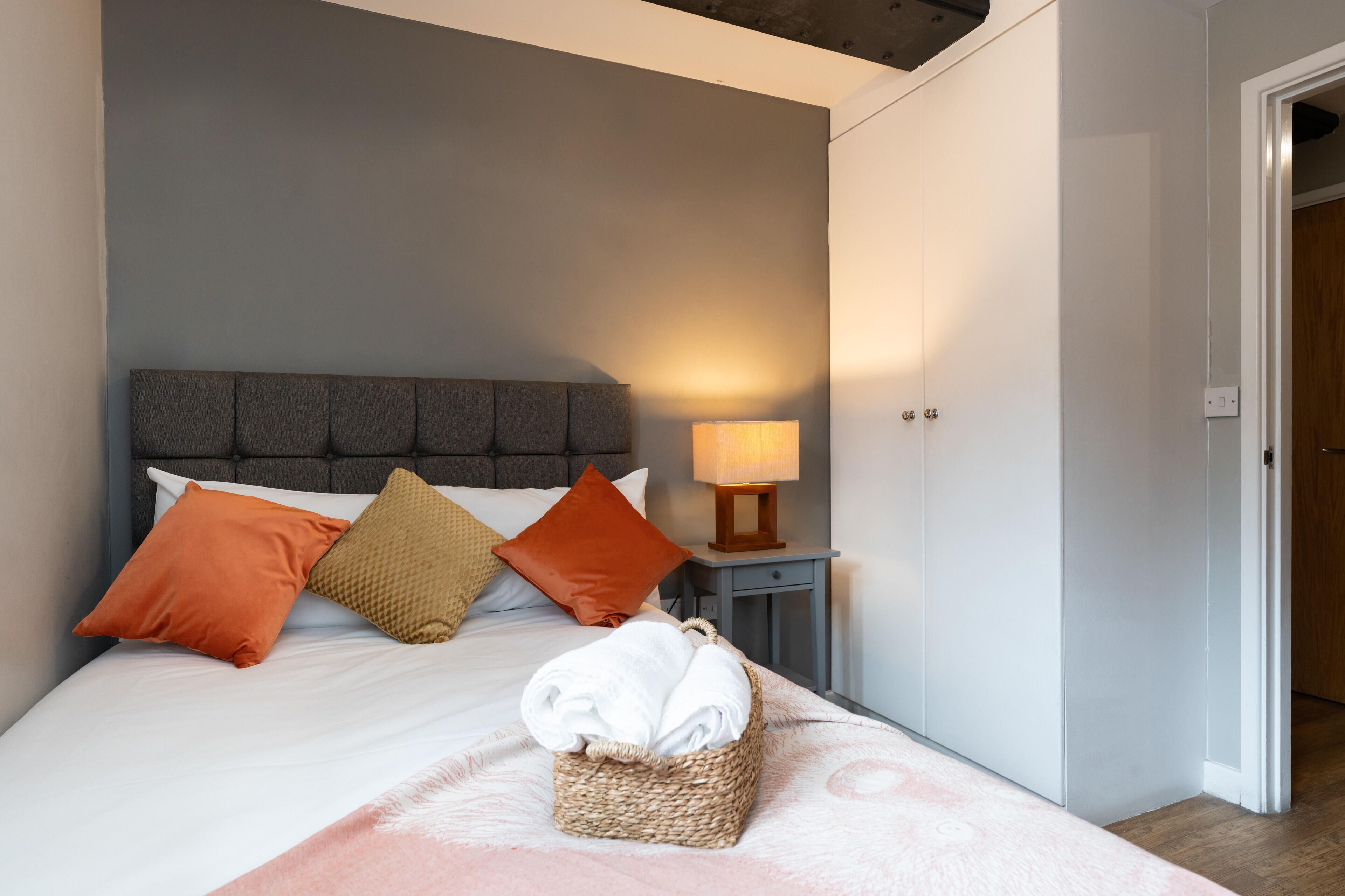 A neatly made bed with a headboard, pillows, a bedside lamp, and a basket of folded towels.