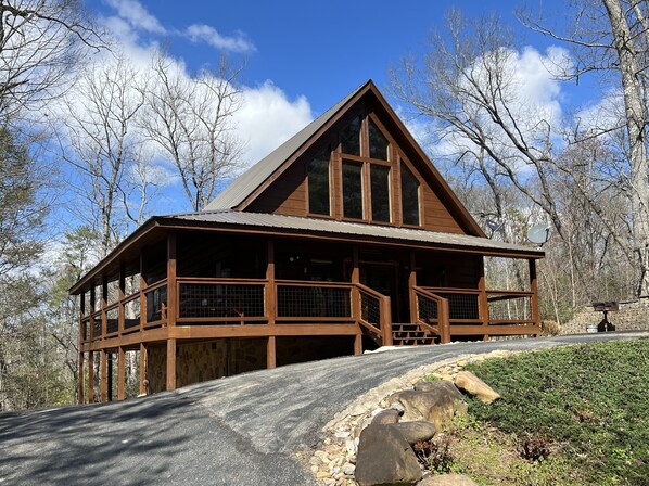 A wooden cabin with a large balcony, surrounded by trees and a clear sky. - Loaded with fun! Hot Tub, Huge Porch, Pool table, Arcade, Starlink, & Outdoor TV (Sevierville)