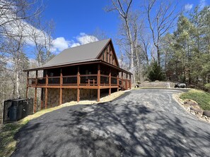 A wooden cabin with a stone foundation, a porch, and a driveway leading to the entrance. - Loaded with fun! Hot Tub, Huge Porch, Pool table, Arcade, Starlink, & Outdoor TV (Sevierville)