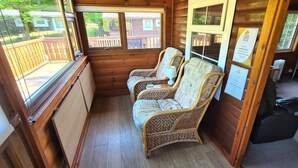 Een blokhut met twee rieten stoelen, een kleine tafel en een raam met uitzicht op een houten terras en bomen.