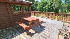 Een houten terras met een picknicktafel en bankjes, omgeven door een houten hek en bomen.