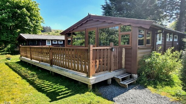 Een houten hut met een terras en een overdekte veranda, omgeven door weelderige begroeiing en een heldere lucht.