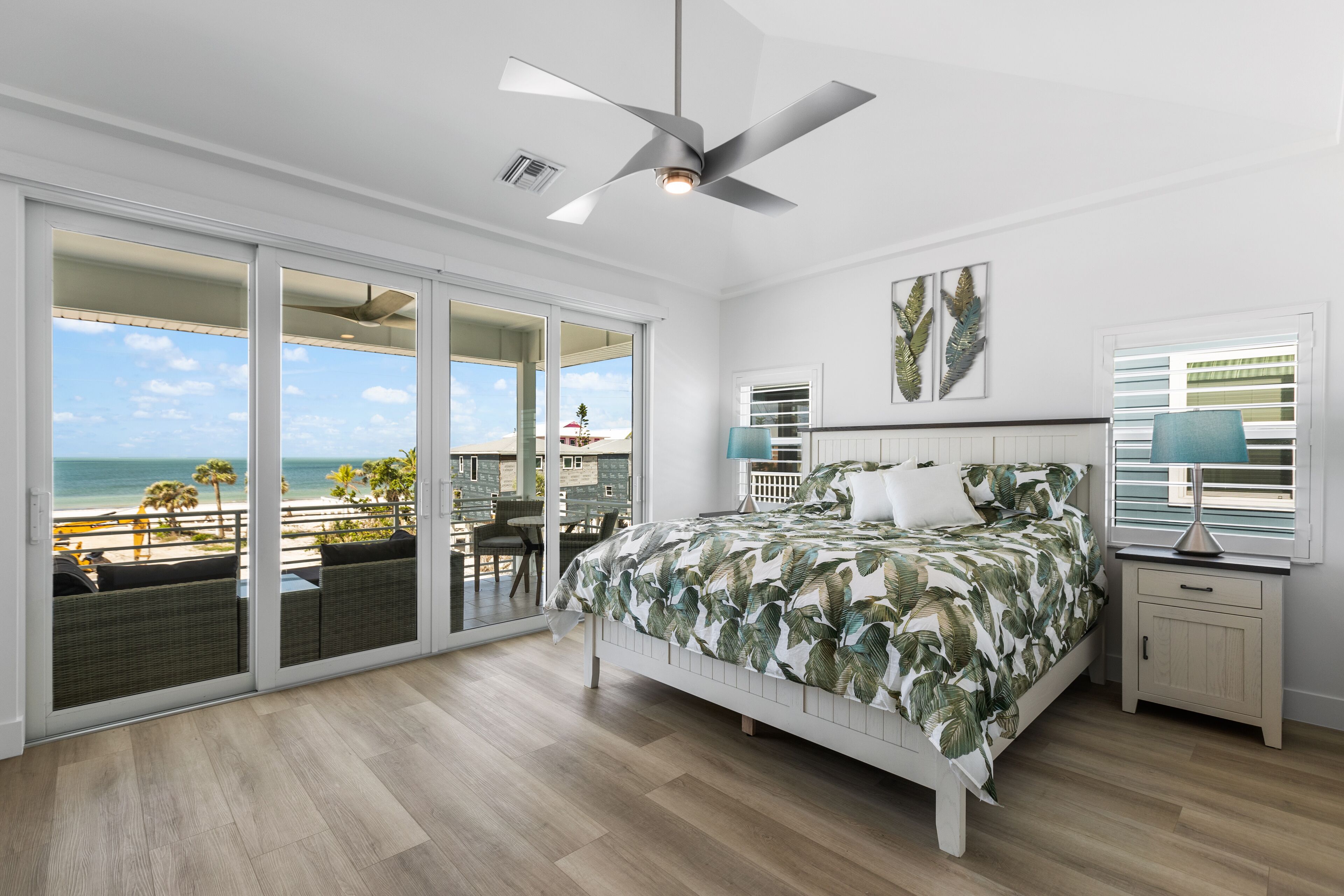 A bedroom with a bed, a ceiling fan, a nightstand with a lamp, and a view of the beach through large sliding glass doors.