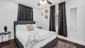 Een moderne slaapkamer met een groot bed, donkere gordijnen, een plafondventilator en decoratieve wandkunst.