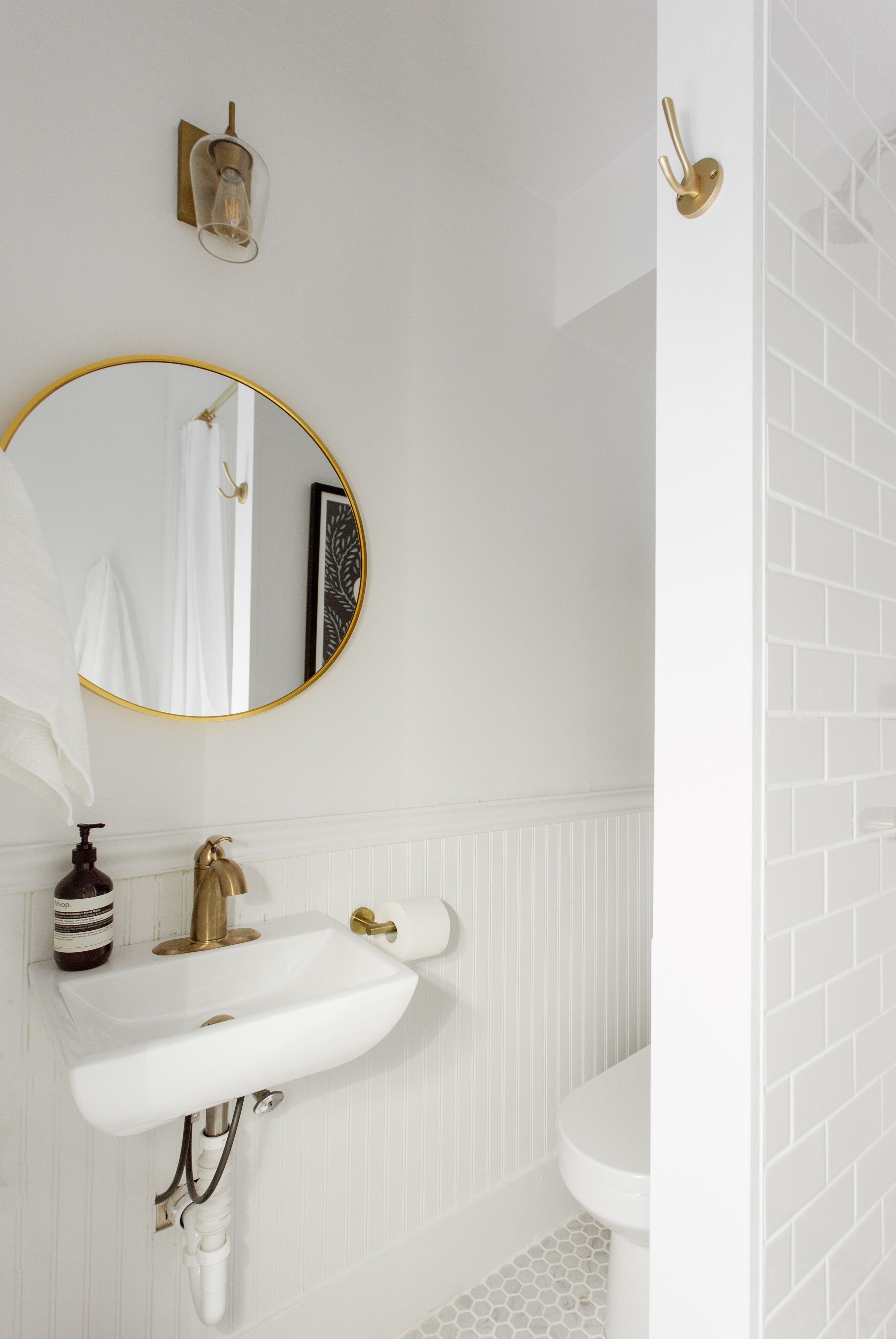 Un baño con lavamanos blanco, detalles dorados, un espejo redondo y un inodoro blanco.