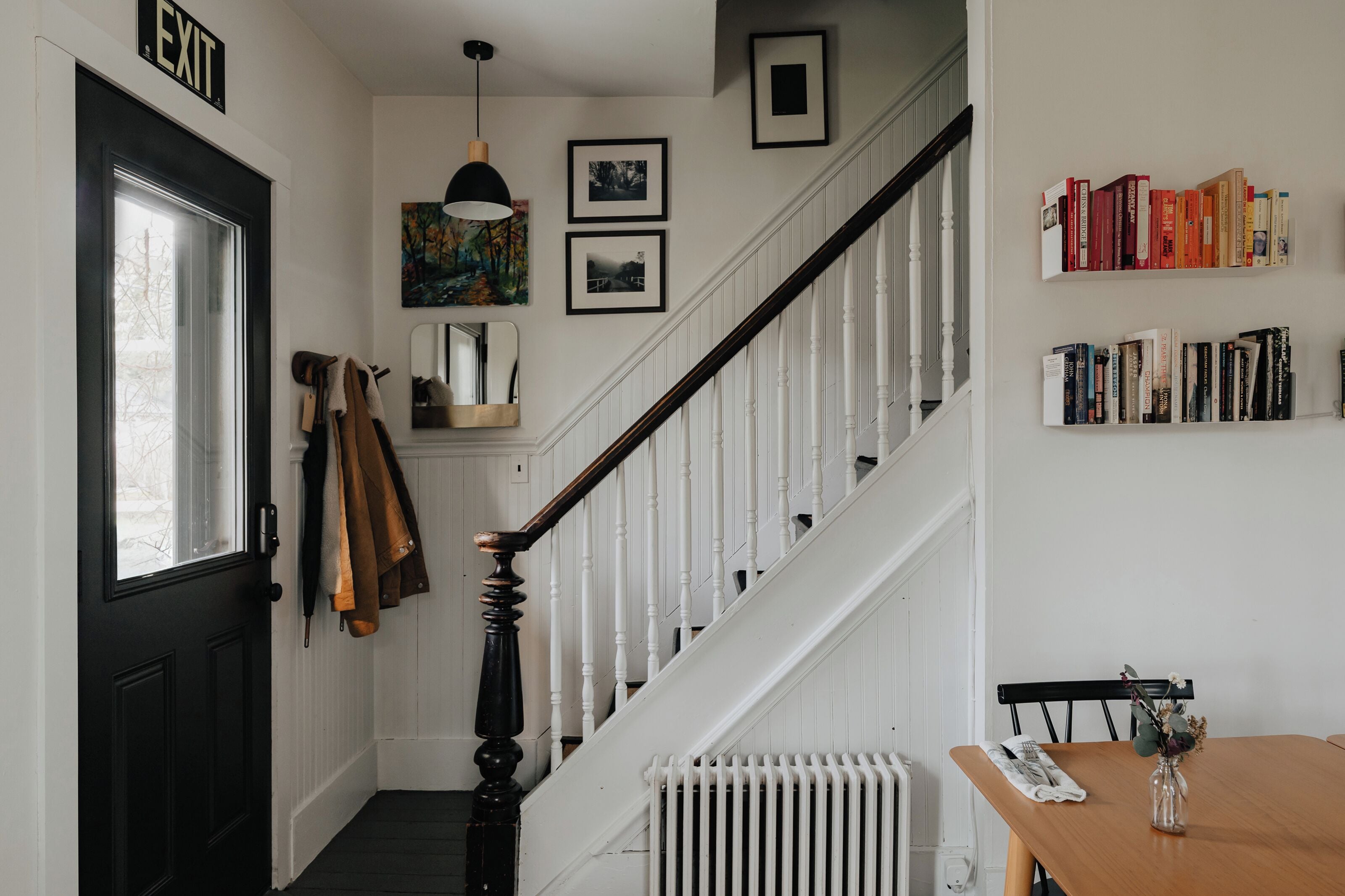 Un escalier avec des rampes blanches et une main courante noire, une porte noire avec une pancarte indiquant « SORTIE », un porte-manteau avec des vestes, un radiateur et une bibliothèque remplie de livres.