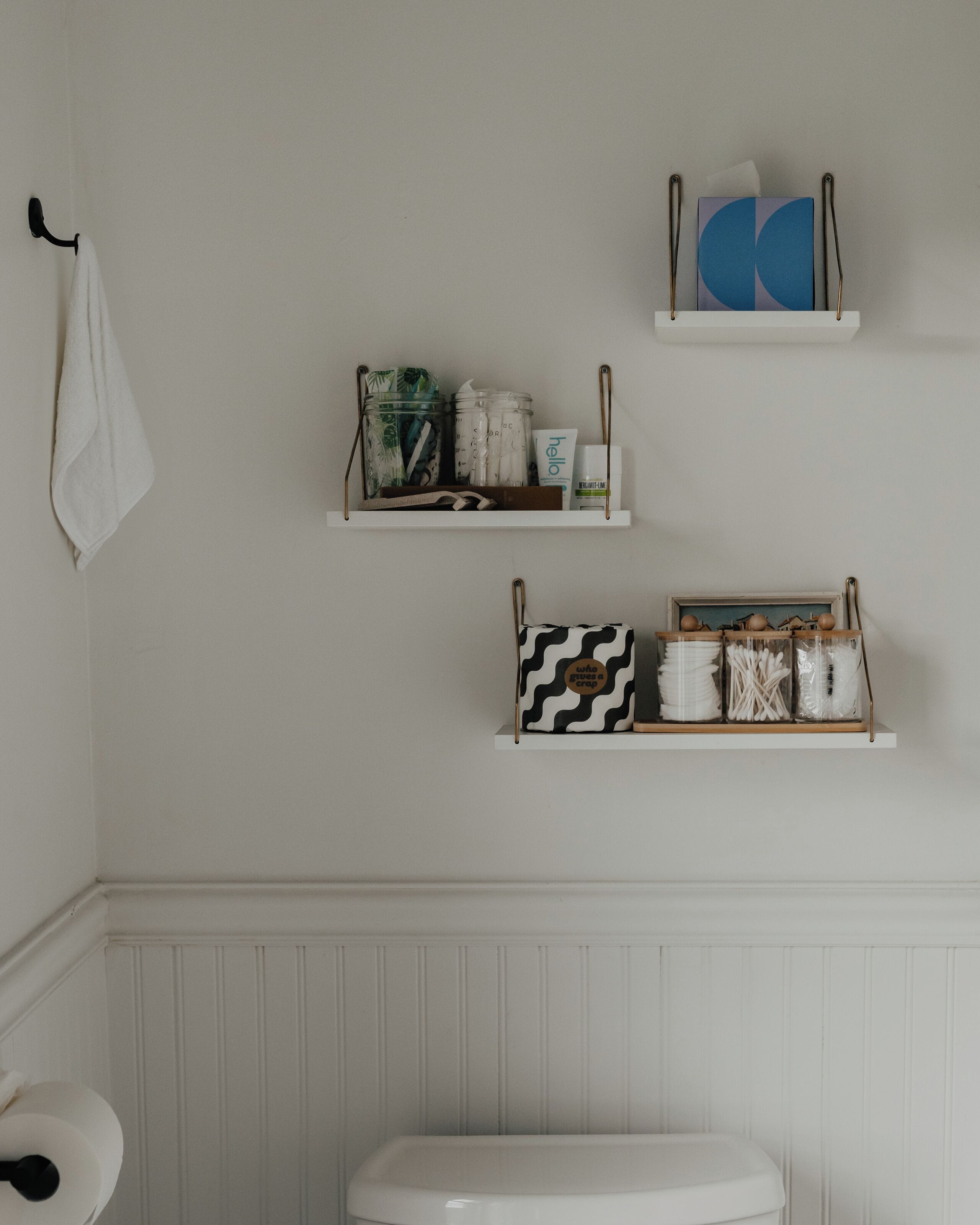 Un baño con inodoro, toallero y tres estantes con artículos de aseo y objetos decorativos.