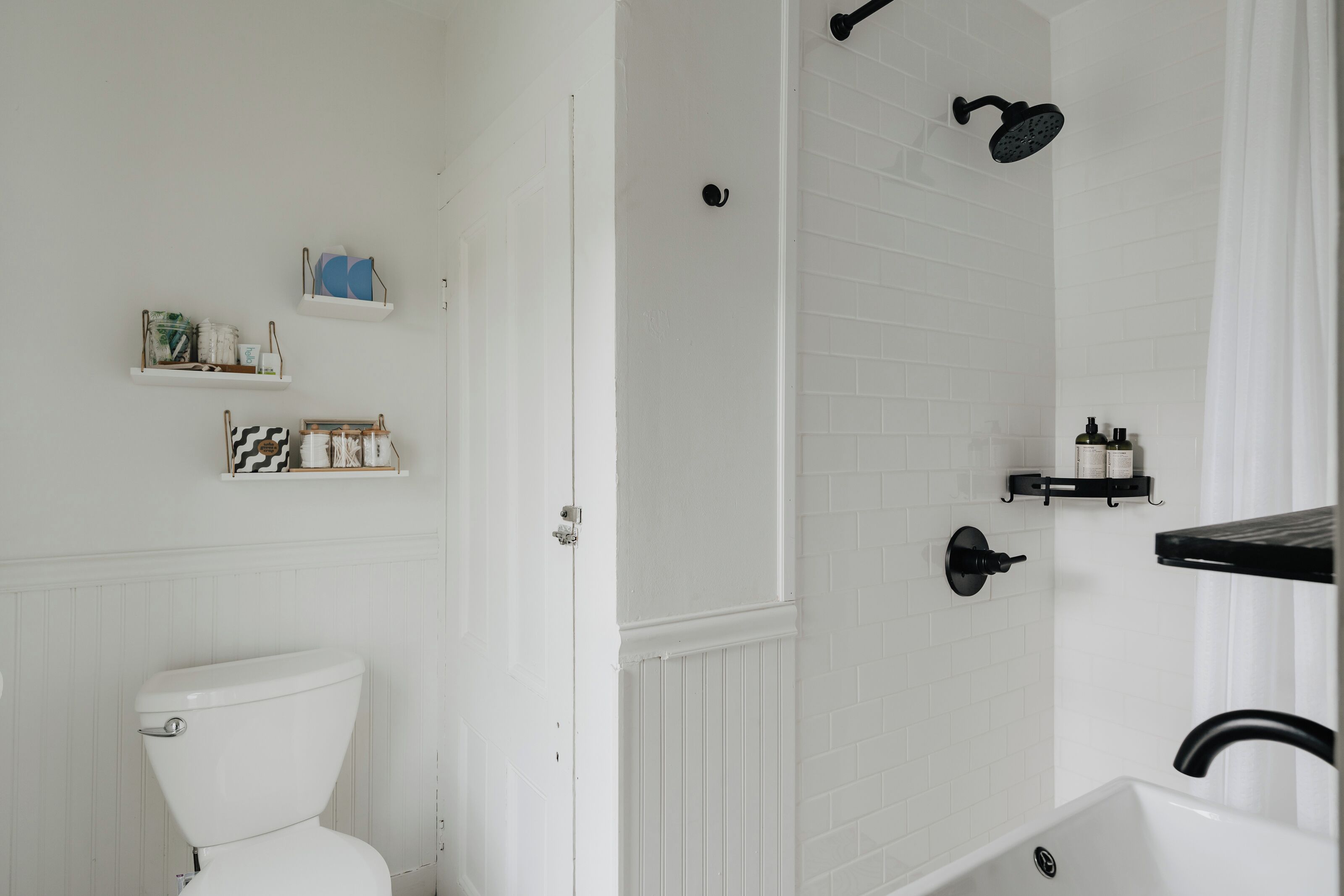 A bathroom with a white toilet, a shower with a black hose, and shelves with toiletries.