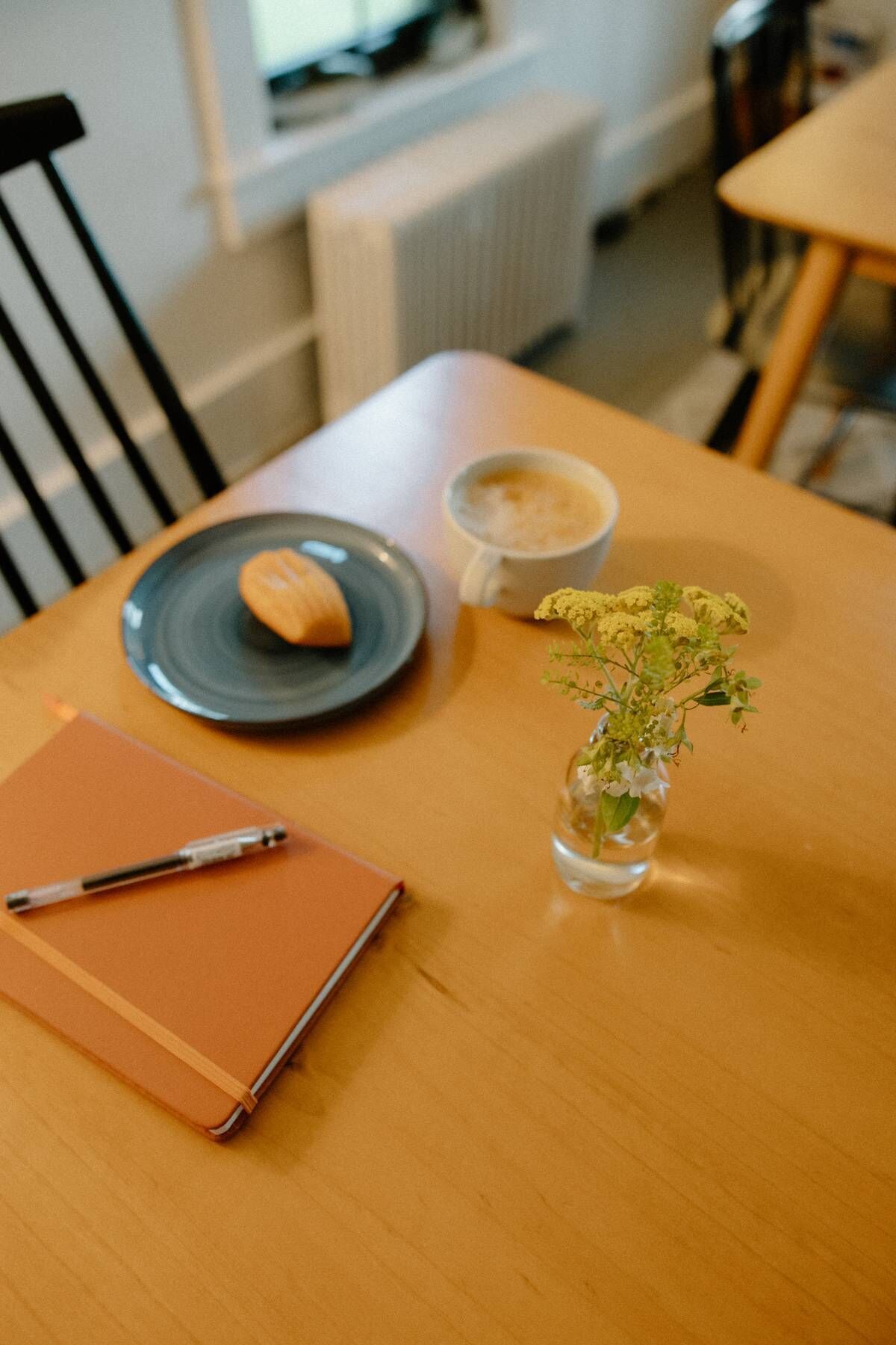 Una mesa de madera con un cuaderno, un bolígrafo, un plato con un pastel, una taza de café y un pequeño florero con flores.