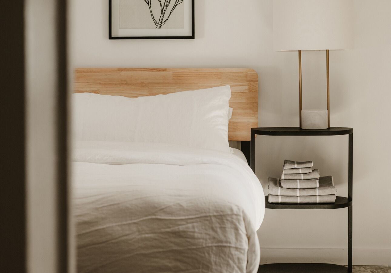 Una cama bien hecha con sábanas blancas, un cabecero de madera, una mesita de noche con una lámpara e impresión enmarcadas de motivos botánicos en la pared.