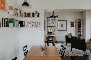 Un comedor con una mesa de madera, sillas y un jarrón con flores. Hay estanterías llenas de libros y una araña.