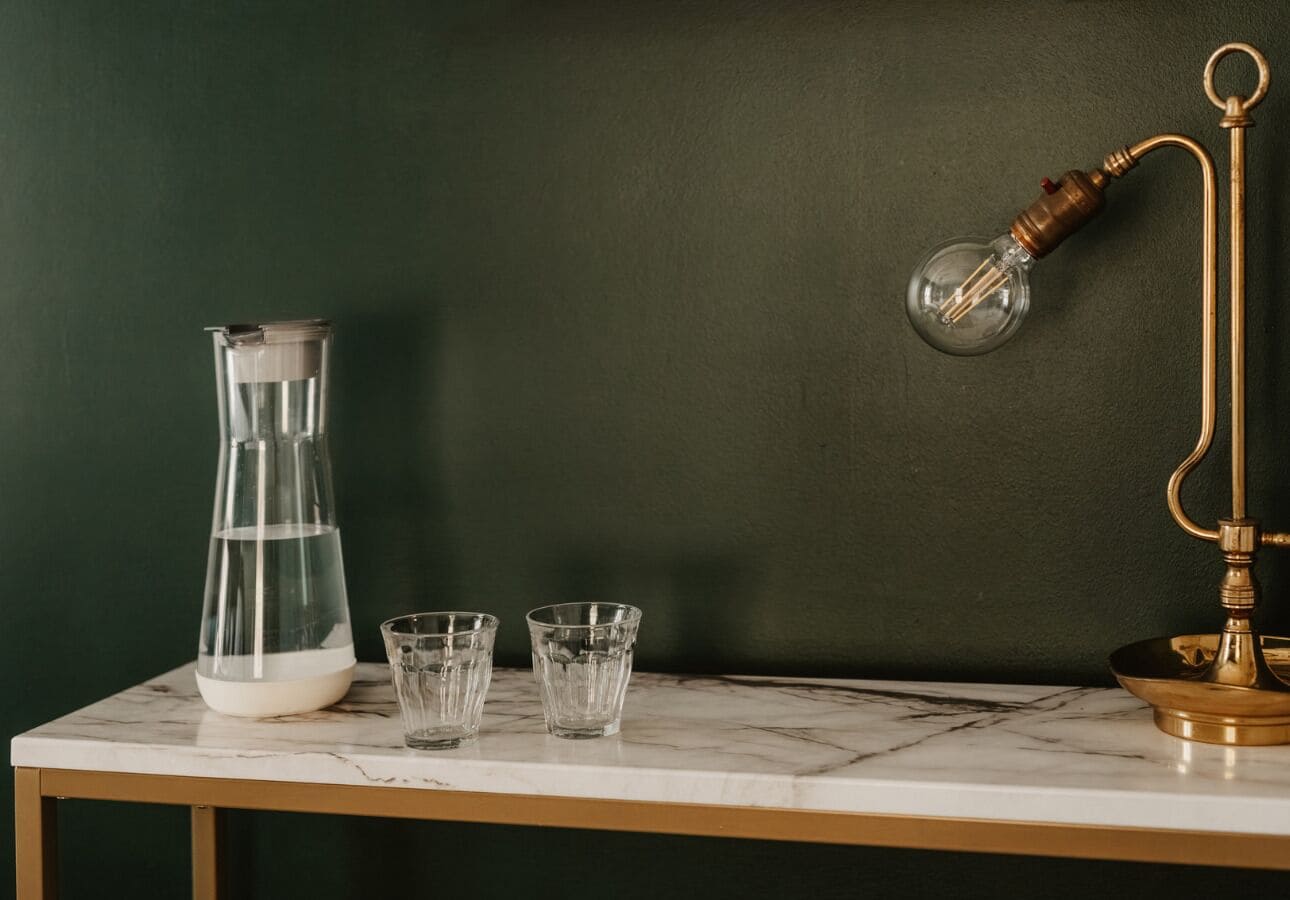 Una mesa de mármol con una jarra de agua, dos vasos y una lámpara de latón frente a una pared oscura.